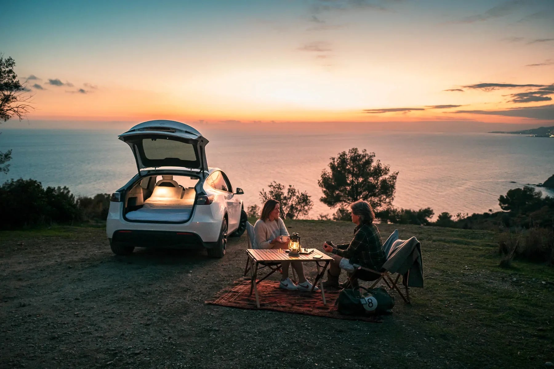 Two campers at a table beside an open Tesla at sunset