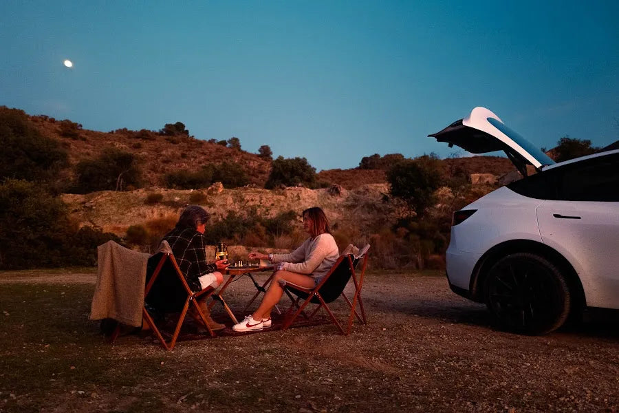 Two campers sitting beside a Tesla Model Y with the hatch open at dusk